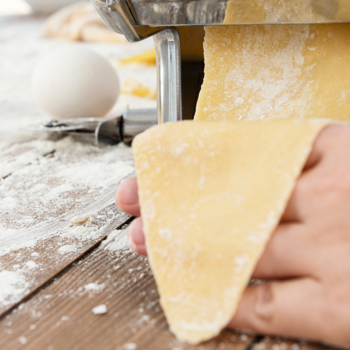 Person using a pasta machine to make pasta on a wooden surface with flour.